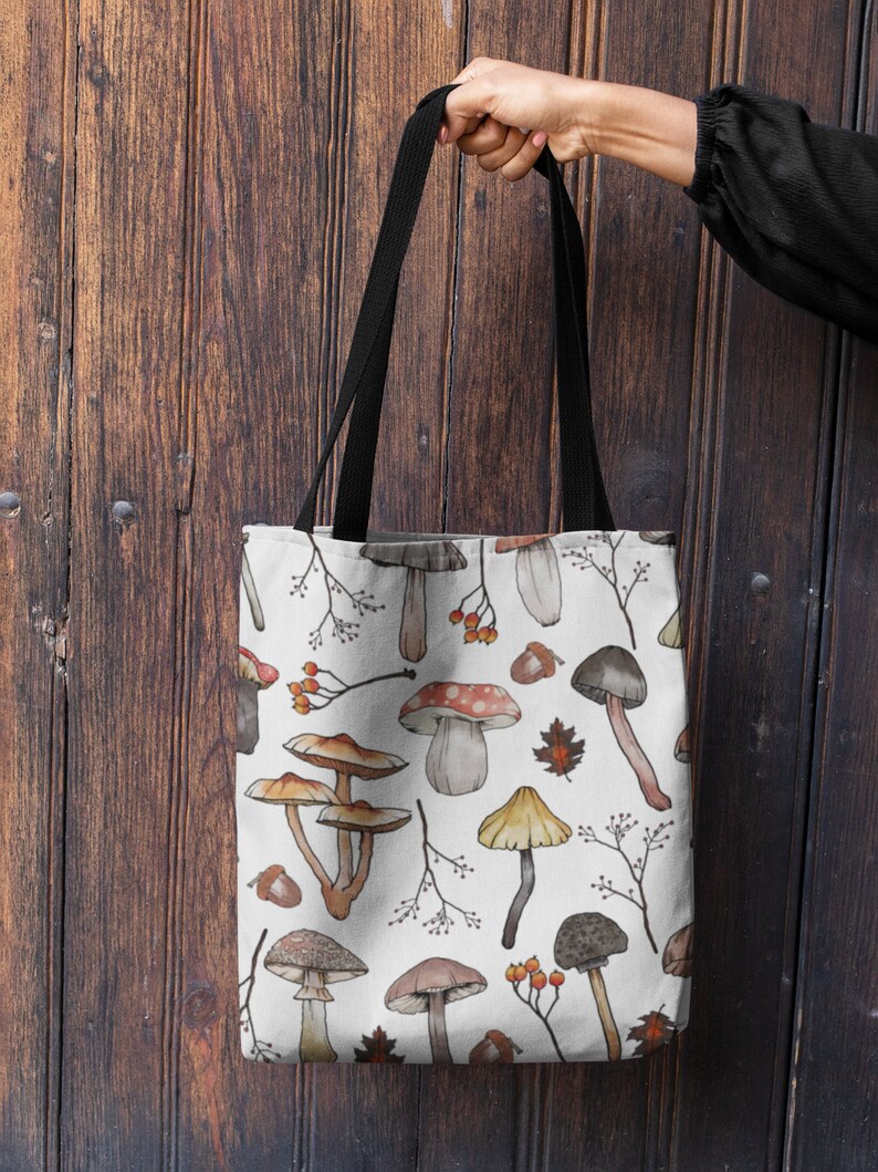 May include: A white canvas tote bag with black straps features a repeating pattern of watercolor mushrooms, leaves, and berries.