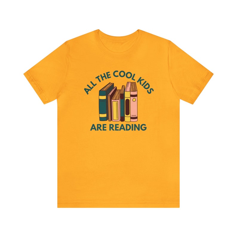 Puede incluir: Una camiseta amarilla con el texto "All the cool kids are reading" y una pila de libros coloridos.