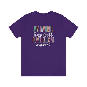 Op de afbeelding: Een paars T-shirt met de tekst "MY FAVORITE baseball PLAYER CALLS ME mom" in kleurrijke letters. Een honkbalafbeelding is onder de tekst opgenomen. Het shirt heeft een ronde hals en korte mouwen.