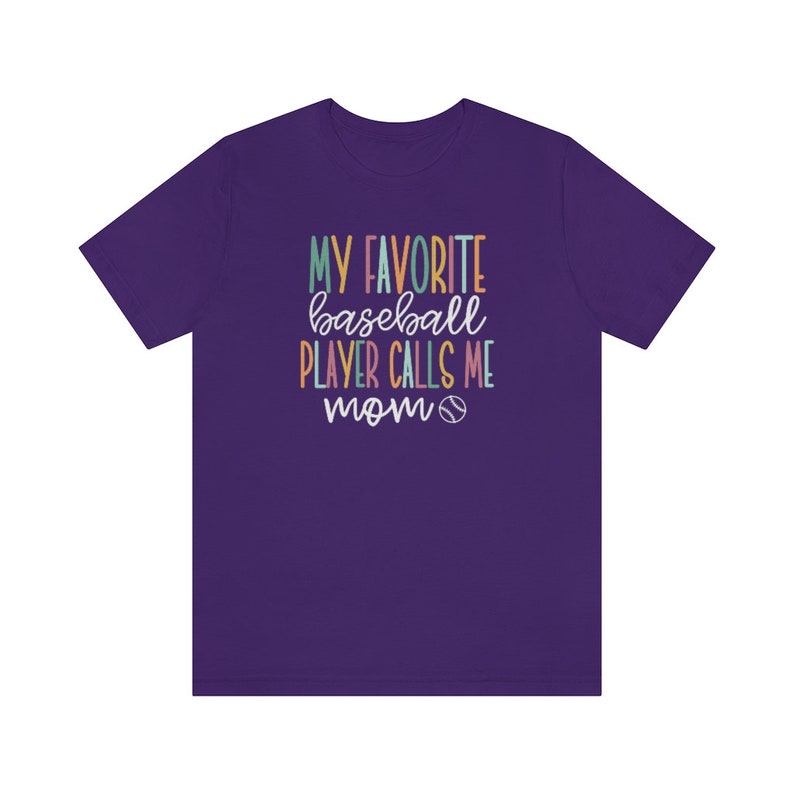 Op de afbeelding: Een paars T-shirt met de tekst "MY FAVORITE baseball PLAYER CALLS ME mom" in kleurrijke letters. Een honkbalafbeelding is onder de tekst opgenomen. Het shirt heeft een ronde hals en korte mouwen.