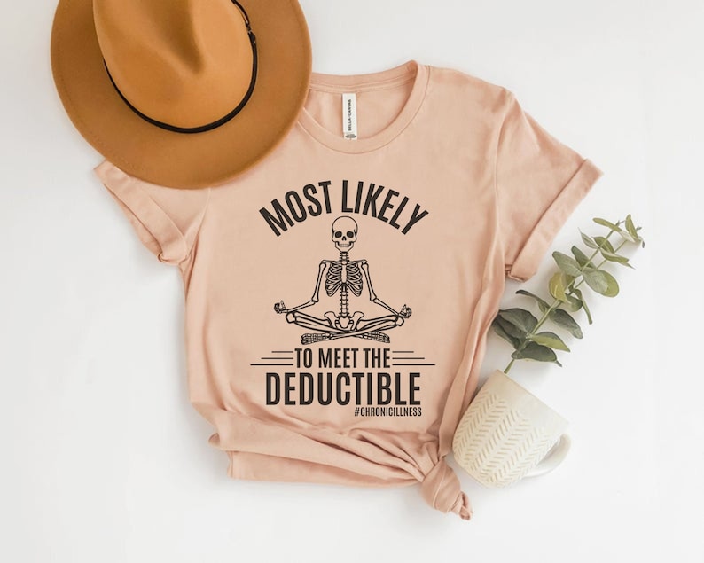 May include: A pale pink t-shirt featuring the words "MOST LIKELY TO MEET THE DEDUCTIBLE" and a meditating skeleton illustration. The hashtag #CHRONICILLNESS is below. A brown hat and a white mug with a plant are in the background.