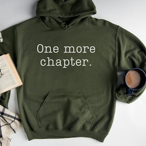 May include: A green hooded sweatshirt with the text "One more chapter." printed in white on the front. The sweatshirt is lying on a white surface with a book, glasses, and a scarf.