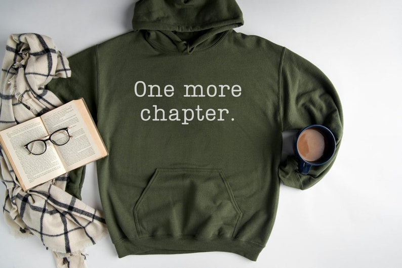 May include: A green hooded sweatshirt with the text "One more chapter." printed in white on the front. The sweatshirt is lying on a white surface with a book, glasses, and a scarf.