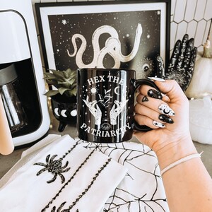 May include: Black ceramic mug with the text "HEX THE PATRIARCHY" and a design of hands, plants, and celestial elements. The mug is held in front of a black and white framed print and a white spiderweb-themed towel.