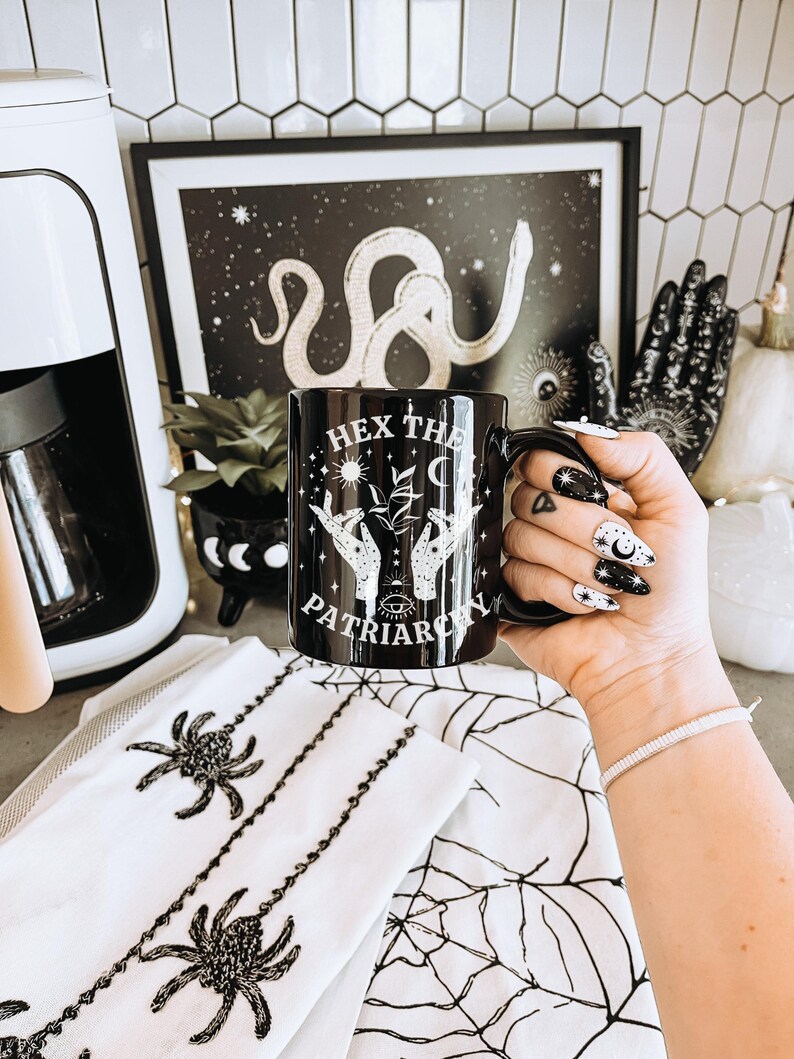 May include: Black ceramic mug with the text "HEX THE PATRIARCHY" and a design of hands, plants, and celestial elements. The mug is held in front of a black and white framed print and a white spiderweb-themed towel.