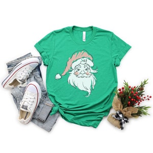 May include: A green t-shirt with a vintage-style illustration of Santa Claus's face. The illustration is in pink, white, and green. The t-shirt is on a white background with a pair of white sneakers and a small bouquet of greenery.