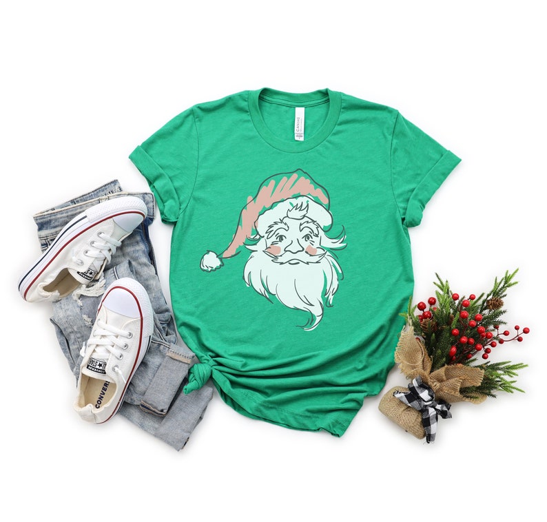 May include: A green t-shirt with a vintage-style illustration of Santa Claus's face. The illustration is in pink, white, and green. The t-shirt is on a white background with a pair of white sneakers and a small bouquet of greenery.