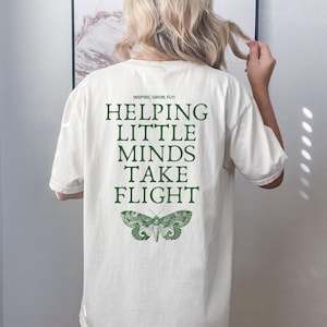 May include: Cream-colored t-shirt with the text "INSPIRE, GROW, FLY! HELPING LITTLE MINDS TAKE FLIGHT" in green. A green moth graphic is below the text. The shirt is a casual, oversized fit.