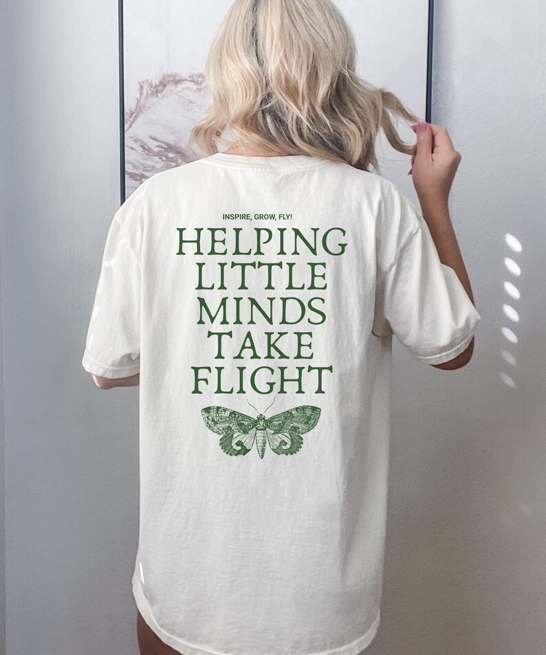 May include: Cream-colored t-shirt with the text "INSPIRE, GROW, FLY! HELPING LITTLE MINDS TAKE FLIGHT" in green. A green moth graphic is below the text. The shirt is a casual, oversized fit.