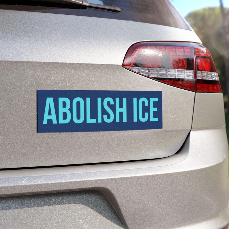 May include: A rectangular blue bumper sticker with the words "ABOLISH ICE" in light blue lettering is affixed to the rear of a silver car. The car's taillight is visible on the right side of the image.
