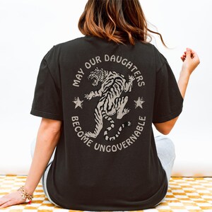 May include: Black t-shirt with a circular graphic on the back featuring a tiger and the text "MAY OUR DAUGHTERS BECOME UNGOVERNABLE". The design includes two stars. The shirt is short-sleeved and has a relaxed fit.