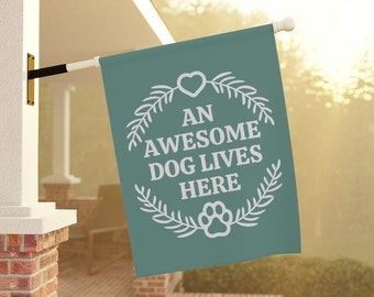 May include: A teal green garden flag with white text and a white paw print. The text reads "An Awesome Dog Lives Here" with a heart and a wreath of leaves surrounding the text.