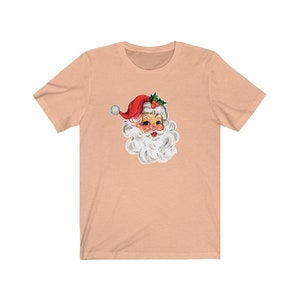May include: A peach-colored t-shirt with a vintage illustration of Santa Claus's face. Santa is wearing a red hat with white trim and a white beard.