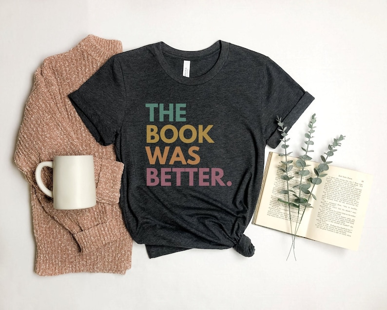 May include: A dark gray t-shirt with the text "THE BOOK WAS BETTER." in colorful letters. The shirt is on a white surface with a book and a sprig of greenery.