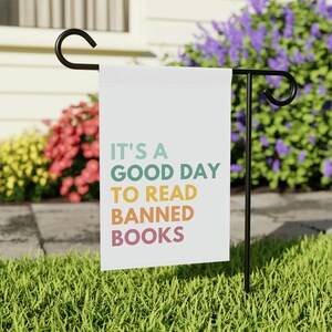 May include: A white garden flag with the text "IT'S A GOOD DAY TO READ BANNED BOOKS" in teal, green, yellow, and orange lettering. The flag is displayed on a black metal stand in a grassy yard with colorful flowers in the background.