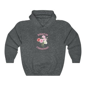 Peut inclure: Un sweat &agrave; capuche gris fonc&eacute; avec le texte "WHAT IN CARNATION?" imprim&eacute; en rose sur le devant. Un bouquet de &oelig;illets roses et blancs est repr&eacute;sent&eacute; &agrave; c&ocirc;t&eacute; du texte.