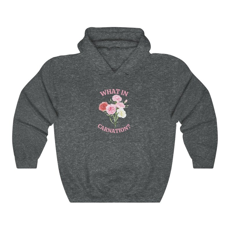 Peut inclure: Un sweat &agrave; capuche gris fonc&eacute; avec le texte "WHAT IN CARNATION?" imprim&eacute; en rose sur le devant. Un bouquet de &oelig;illets roses et blancs est repr&eacute;sent&eacute; &agrave; c&ocirc;t&eacute; du texte.