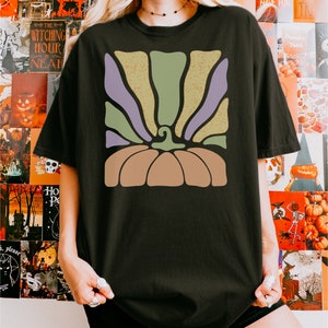 May include: A black t-shirt featuring a graphic print of a stylized pumpkin. The pumpkin design consists of a solid tan base with a curved stem and radiating vertical stripes in alternating shades of muted sage green and light purple.