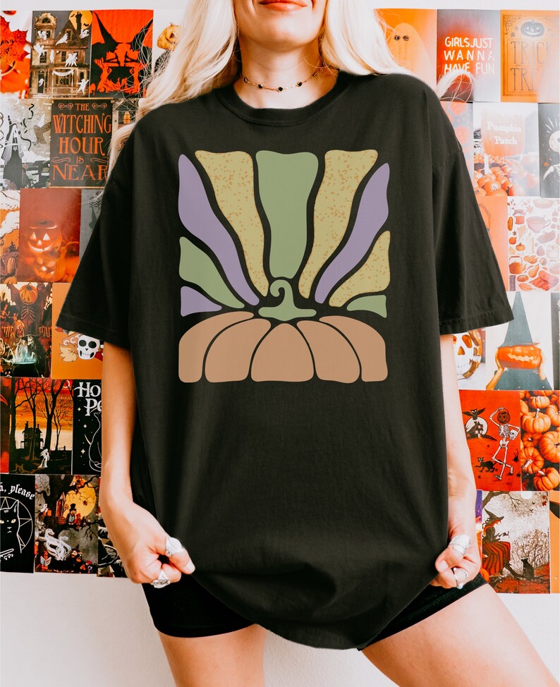 May include: A black t-shirt featuring a graphic print of a stylized pumpkin. The pumpkin design consists of a solid tan base with a curved stem and radiating vertical stripes in alternating shades of muted sage green and light purple.