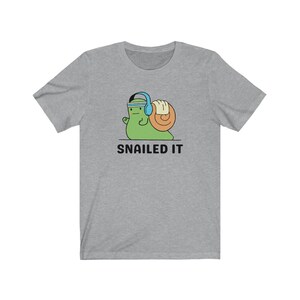 K&ouml;nnte beinhalten: Ein graues T-Shirt mit einer Cartoon-Schnecke, die Kopfh&ouml;rer und ein Stirnband tr&auml;gt. Die Schnecke hebt ein Gewicht. Der Text "SNAILED IT" ist unter der Schnecke gedruckt.