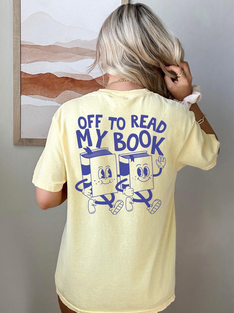 May include: Yellow t-shirt with the text "OFF TO READ MY BOOK" in blue. The shirt features a graphic of two animated books with arms, legs, and smiling faces. The shirt is a light yellow color.