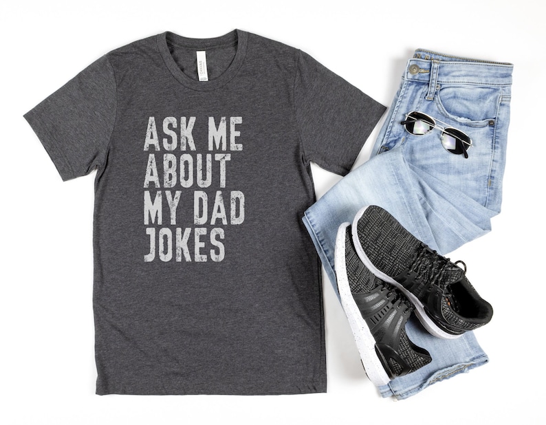 K&ouml;nnte beinhalten: Ein dunkelgraues T-Shirt mit dem Aufdruck "Ask me about my dad jokes" in wei&szlig;. Das Shirt liegt flach auf einer wei&szlig;en Oberfl&auml;che mit einer blauen Jeans und schwarzen Turnschuhen.