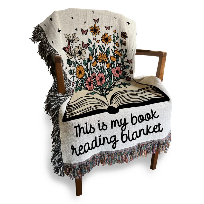 Peut inclure: Une couverture en tissu tiss&eacute; de couleur cr&egrave;me avec un motif de livre et de fleurs. Le livre est ouvert avec des fleurs qui en sortent. Le texte sur la couverture est "This is my book reading blanket".