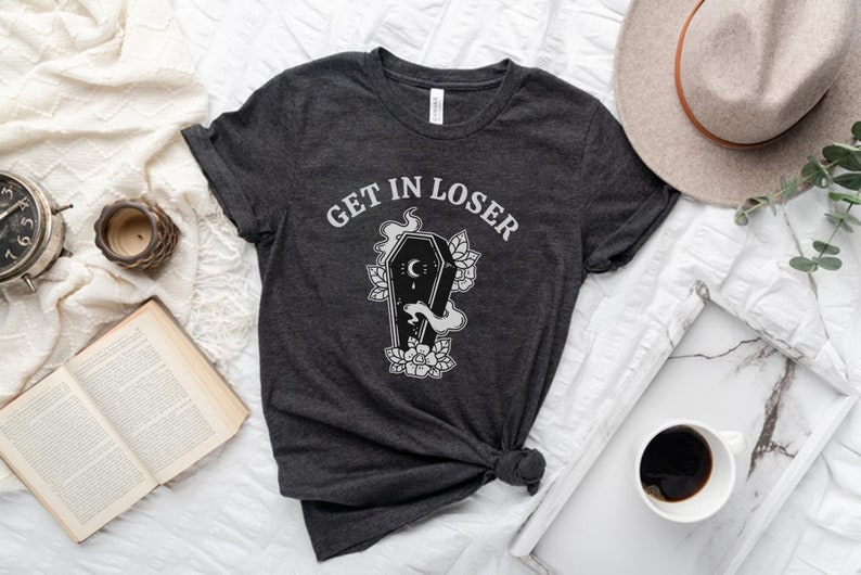 May include: A dark grey t-shirt with the text "GET IN LOSER" printed in white. The shirt features a black and white illustration of a coffin with a crescent moon and flowers.