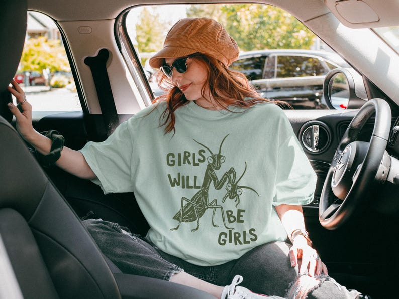 May include: A light green t-shirt with the text "GIRLS WILL BE GIRLS" and a graphic of praying mantises. The person in the image is wearing a brown corduroy hat and sunglasses. The image is taken inside a car.