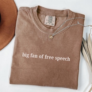 May include: A brown Comfort Colors t-shirt with the text "big fan of free speech" in white. A silver necklace with a pendant is draped across the shirt. A brown hat and dried plant are in the background.