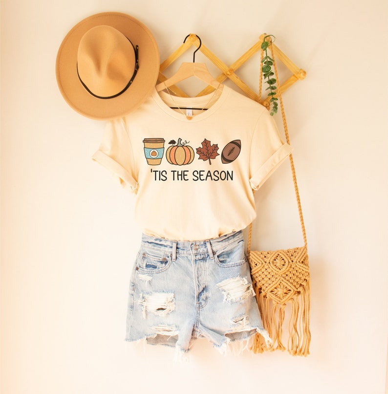 May include: A beige t-shirt with a graphic of a coffee cup, pumpkin, leaf, and football with the text 'Tis the season'. The shirt is hanging on a wooden hanger with a brown hat and a woven bag.