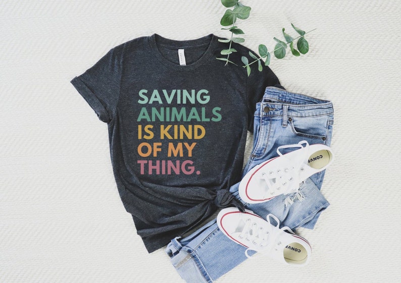 Puede incluir: Camiseta gris oscuro con el texto "SAVING ANIMALS IS KIND OF MY THING" en letras coloridas. Se combina con vaqueros azules y zapatillas blancas. Una ramita verde a&ntilde;ade un toque natural al conjunto casual.