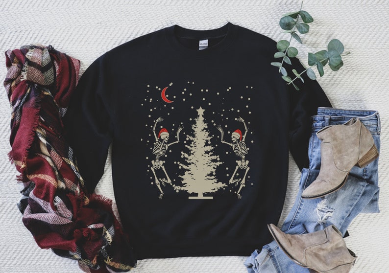 May include: Black sweatshirt with a Christmas tree and two skeletons wearing Santa hats dancing around it. The skeletons are white and the tree is beige. There is a crescent moon and falling snow in the background.