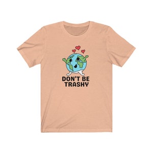 May include: Peach-coloured t-shirt with a cartoon Earth character, arms, legs, and three red hearts. The text "DON'T BE TRASHY" is printed in black below the Earth illustration. The shirt is made of a soft material.