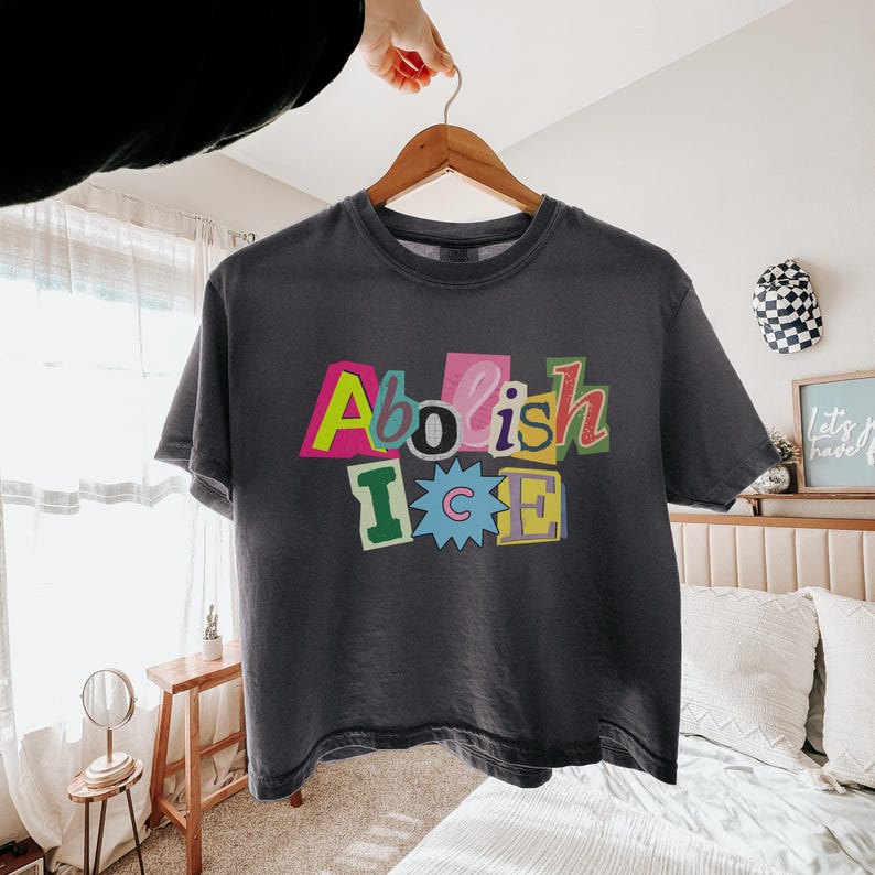 May include: Dark gray t-shirt with the words "Abolish ICE" in colorful, cut-out style letters. The shirt is hanging on a wooden hanger, with a hand holding the hanger. The shirt is a casual, unisex style.