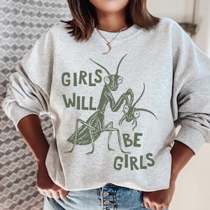 May include: A light gray sweatshirt with the text "GIRLS WILL BE GIRLS" in green. The sweatshirt features a green illustration of a praying mantis and a smaller mantis.