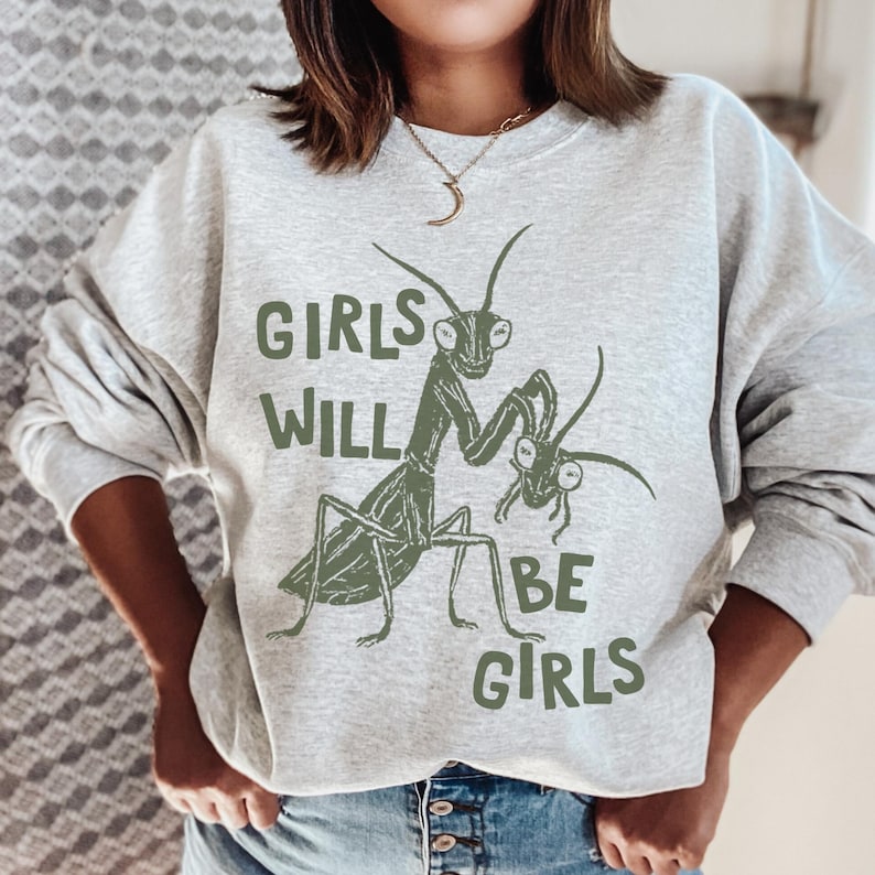 May include: A light gray sweatshirt with the text "GIRLS WILL BE GIRLS" in green. The sweatshirt features a green illustration of a praying mantis and a smaller mantis.