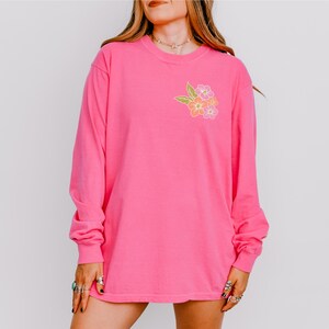 Puede incluir: Una camiseta rosa de manga larga con un dise&ntilde;o floral en el pecho izquierdo. Las flores son amarillas, naranjas y rosas.