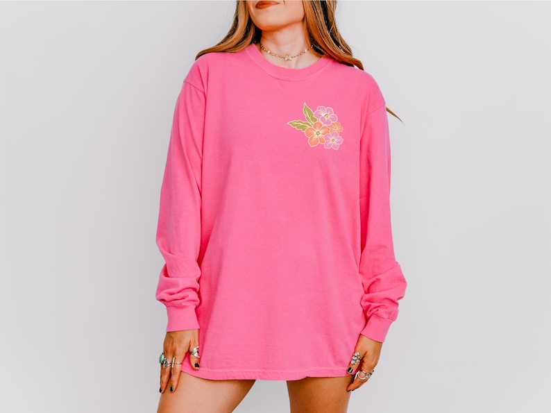 Puede incluir: Una camiseta rosa de manga larga con un dise&ntilde;o floral en el pecho izquierdo. Las flores son amarillas, naranjas y rosas.