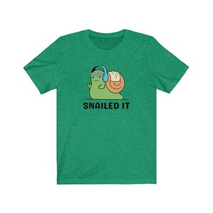 K&ouml;nnte beinhalten: Gr&uuml;nes T-Shirt mit einer Cartoon-Schnecke, die Kopfh&ouml;rer tr&auml;gt und ein Gewicht hebt. Der Text "SNAILED IT" ist unter der Schnecke gedruckt.