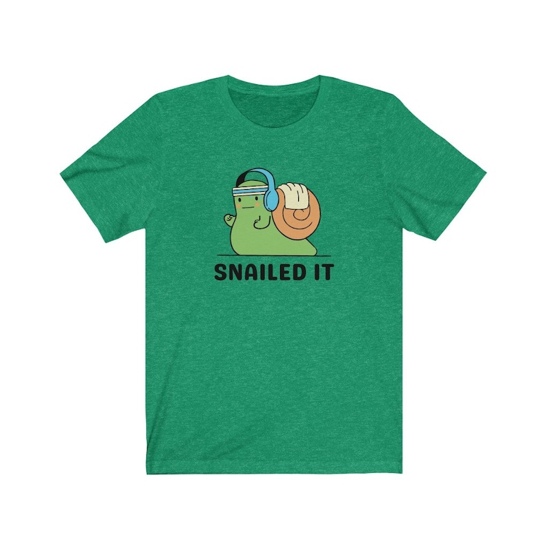 K&ouml;nnte beinhalten: Gr&uuml;nes T-Shirt mit einer Cartoon-Schnecke, die Kopfh&ouml;rer tr&auml;gt und ein Gewicht hebt. Der Text "SNAILED IT" ist unter der Schnecke gedruckt.