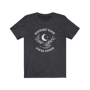 May include: Dark heather grey t-shirt with a white graphic design featuring a crescent moon, stars, and two hands holding a sprig of leaves. The text "Support Your Local Coven" is printed below the graphic.