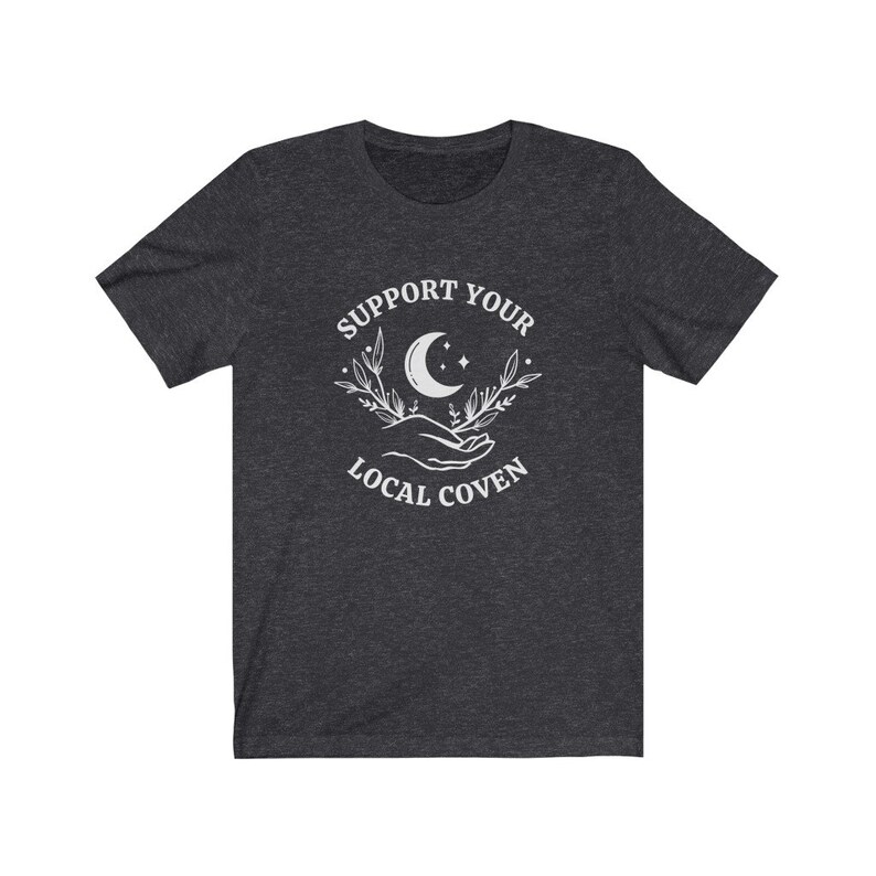 May include: Dark heather grey t-shirt with a white graphic design featuring a crescent moon, stars, and two hands holding a sprig of leaves. The text "Support Your Local Coven" is printed below the graphic.