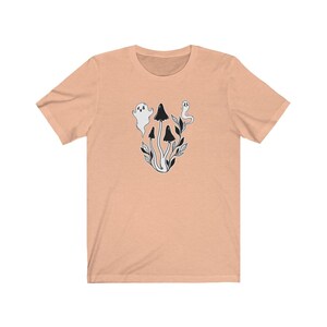 Puede incluir: Una camiseta de color melocot&oacute;n con una ilustraci&oacute;n en blanco y negro de setas y fantasmas. La ilustraci&oacute;n representa tres setas con tallos blancos y sombreros negros, y dos fantasmas con cuerpos blancos y ojos negros.