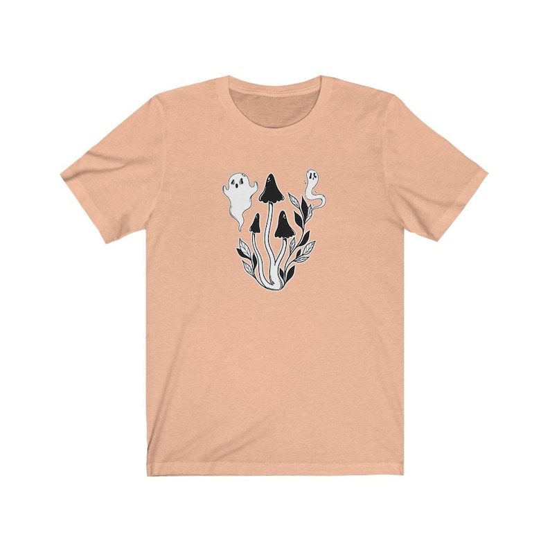 Puede incluir: Una camiseta de color melocot&oacute;n con una ilustraci&oacute;n en blanco y negro de setas y fantasmas. La ilustraci&oacute;n representa tres setas con tallos blancos y sombreros negros, y dos fantasmas con cuerpos blancos y ojos negros.