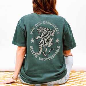 May include: Dark green t-shirt with a circular graphic on the back. The graphic features a tiger illustration and the text "MAY OUR DAUGHTERS BECOME UNGOVERNABLE". The shirt is a relaxed fit.