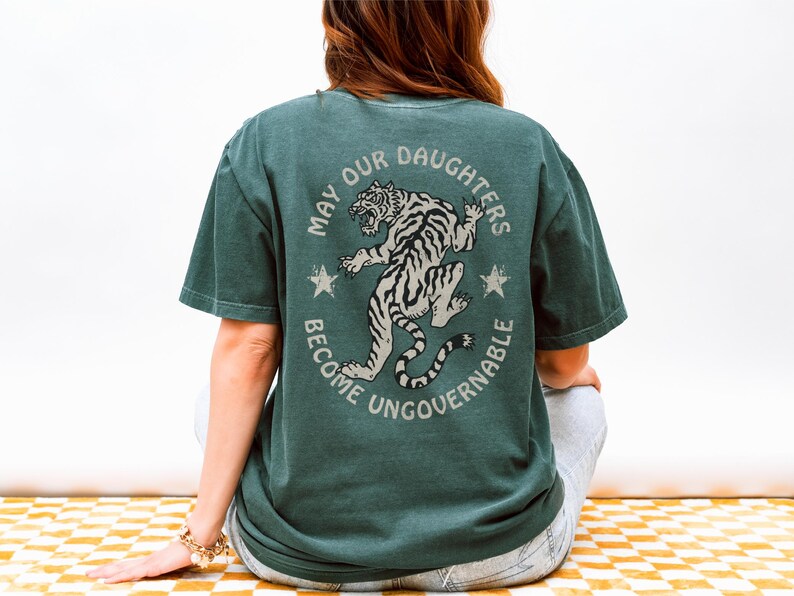 May include: Dark green t-shirt with a circular graphic on the back. The graphic features a tiger illustration and the text "MAY OUR DAUGHTERS BECOME UNGOVERNABLE". The shirt is a relaxed fit.