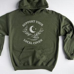 May include: A green hooded sweatshirt with the text "Support Your Local Coven" and a white graphic of a crescent moon, stars, and hands holding a sprig of leaves.