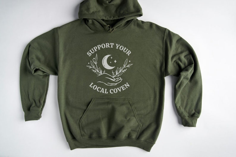 May include: A green hooded sweatshirt with the text "Support Your Local Coven" and a white graphic of a crescent moon, stars, and hands holding a sprig of leaves.