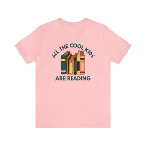 Puede incluir: Camiseta rosa con un gr&aacute;fico de una pila de libros y el texto "All the cool kids are reading."